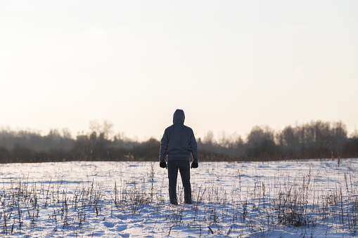 Young adult man standing alone on white snowy field and looking far away. Cold winter day. Back view. Empty place for text, quote or sayings on light sky background.