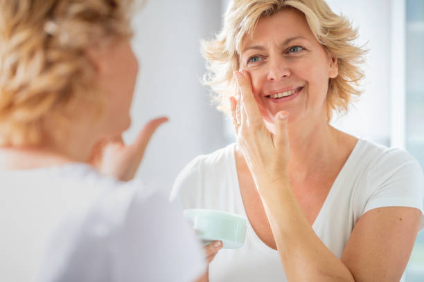 cheerful woman putting on facial cream in front of the mirror - moisturizer women cosmetics body imagens e fotografias de stock