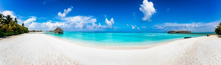 Wide panoramic view of a tropical beach with turquoise sea, Palm trees and water lodges over the ocean in the Maldives islands