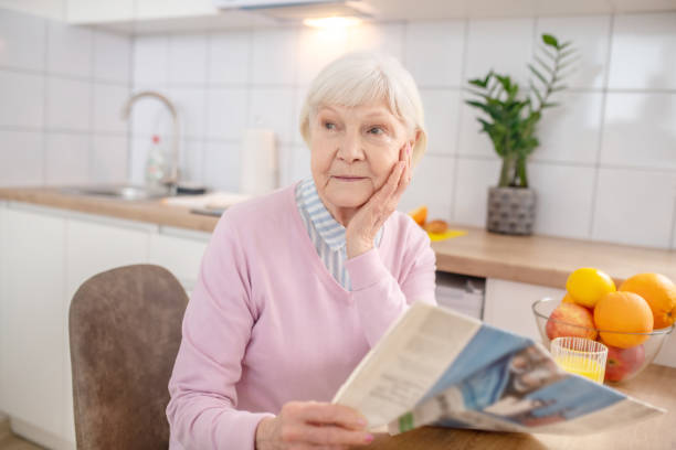 istock Elderly grey-haired woman reading a newspaper in the kitchen 1297175344
