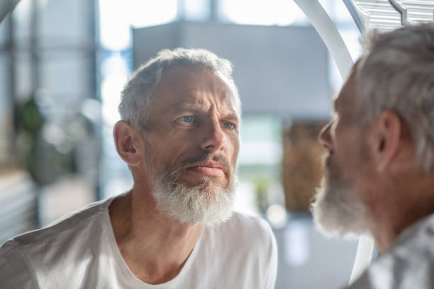 Bearded grey-haired man looking at the mirror Looking at the reflection. A bearded grey-haired man looking at the mirror mesmerised stock pictures, royalty-free photos & images