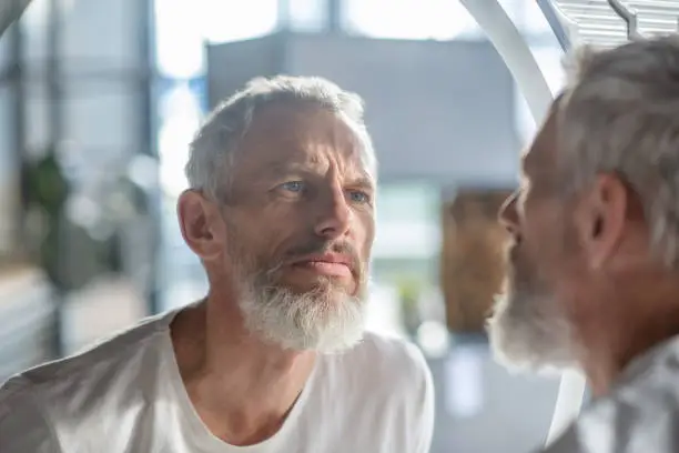 Photo of Bearded grey-haired man looking at the mirror