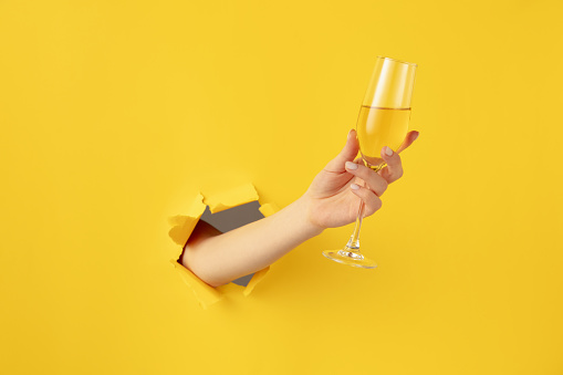 Beautiful woman girl hand with champagne flute glass breaks through yellow paper background. Copy space for ad, design, text. Concept of holidays, celebrations and greeting.