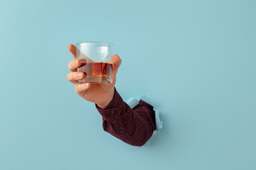 Strong alcohol. Man's hand holding glass for whiskey and breaking through blue paper background. Copy space for ad, design, text. Concept of holidays, traditions, celebrations and greeting. Faceless.