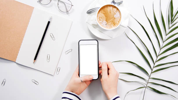 istock Flatlay on white work space desk with woman's hands holding white smartphone with white copyspace 1297183719