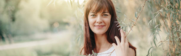 Beautiful happy smiling middle age young Caucasian woman with long dark brown red hair. Casual style real woman female in white dress outdoors in park nature. Web banner header. Beautiful happy smiling middle age young Caucasian woman with long dark brown red hair. Casual style real woman female in white dress outdoor in park nature. Web banner header. willow tree photos stock pictures, royalty-free photos & images