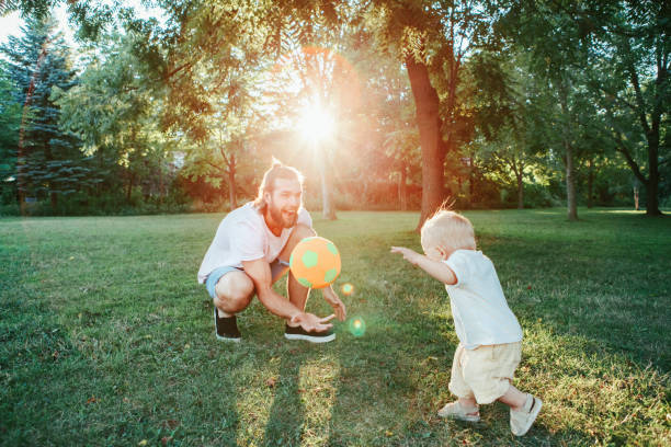 день отцов. отец играет в мяч с малышом мальчик на открытом воздухе. родитель проводит время вместе с ребенком сына в парке. аутентичный обр� - action family photograph fathers day стоковые фото и изображения