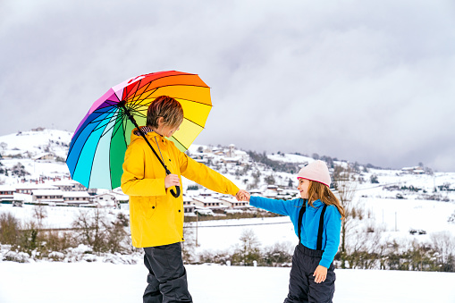 woman and daughter happily holding hands carries a multicolored umbrella in the snow on a mountain with a beautiful landscape and wears a yellow jacket and the girl a blue sweater and pink hat