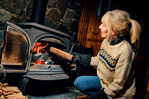 Woman putting a well dried log into a glowing wood burning stove.