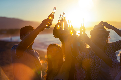 Group of young people partying on the beach at sunset. They are all holding bottles of beer and celebrating with a toast. Silhouette with the beach, sun and sea in the background