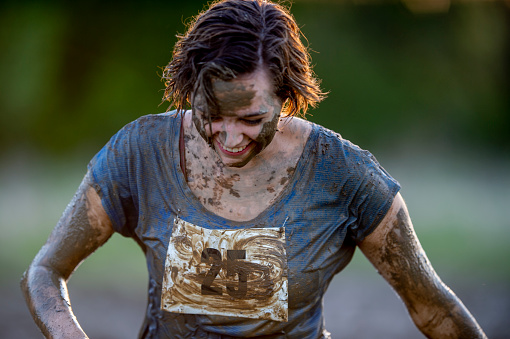 A young female adult of Caucasian ethnicity is wearing sports clothing outdoors and is covered in mud. She is participating in a non-profit mud run competition.