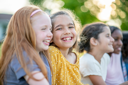 An elementary age girl of mixed race ethnicity is hanging out with her friends at the park outdoors. She is sitting next to another female friend who is of Caucasian ethnicity. Her other friends can be seen in the background.