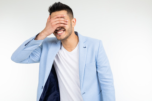 guy in a blue jacket covered his face with his hands and laughs on a white studio background.
