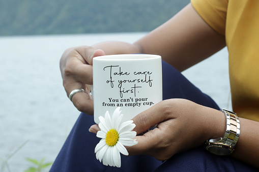 Inspirational quote - Take care of yourself first. You cannot pour from an empty cup. With young woman sitting in the lake holding cup of tea or coffee and a flower. Self love and care concept.
