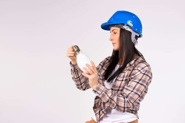 istock A female electrician in a blue hard hat examines an LED lamp on a white background with empty side space. 1297210119
