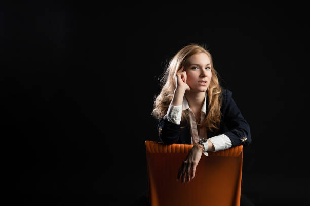istock sitting on an orange chair. Smiling looking at camera, webinar activity concept, on black background in studio, in blazer business woman, blonde 1297215900