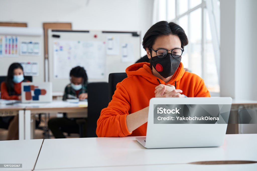 Smart guy is sitting at the table opposite a laptop and looking at the screen Smart guy is sitting at the table opposite a laptop and looking at the screen with bored emotions. Man studying at the classroom during pandemic. Social distancing concept Learning Stock Photo