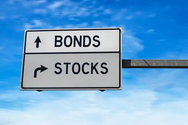 Photo of Road sign with words bonds and stocks. White two street signs with arrow on metal pole on blue sky background.