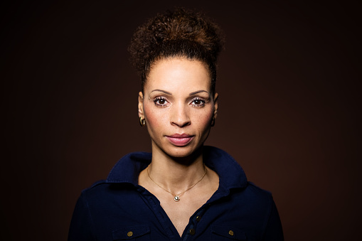 Portrait of a confident mid adult woman staring at camera. Attractive african woman wearing blue shirt with curly hair against brown background.