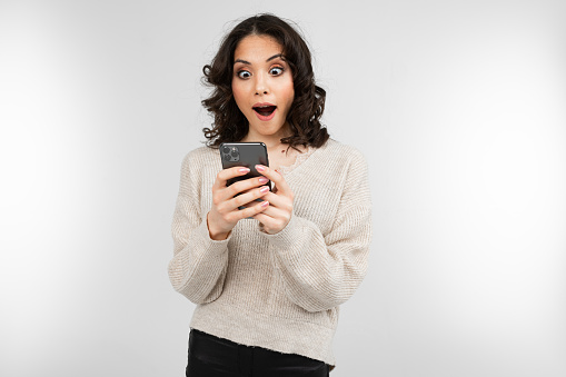 surprised brunette girl in a white sweater with a phone in his hand on a white background with copy space.