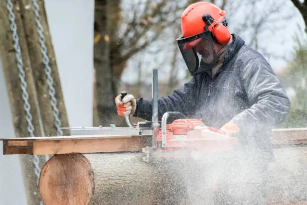 Man cutting log with a chainsaw for making furniture. He is a carpenter preparing wood for his work. He is wearing protective clothing, helmet, ear protection and gloves. The photograph is taken outdoor.
