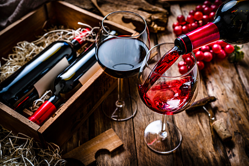 High angle view of a bottle pouring red wine into a wineglass shot on rustic wooden table. A second wineglass filled with red wine, a box with two new wine bottles, straw, a corkscrew with a cork and grapes complete the composition. Predominant colors are red and brown. High resolution 42Mp studio digital capture taken with SONY A7rII and Zeiss Batis 40mm F2.0 CF lens