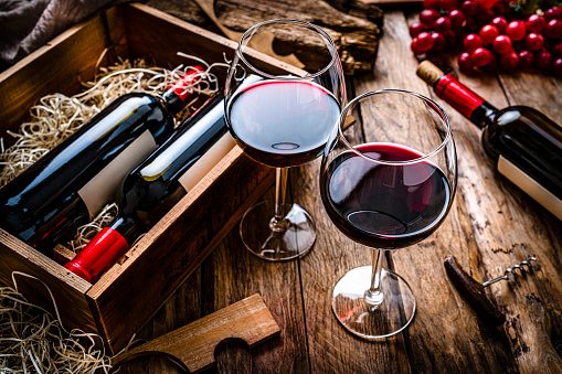 High angle view of two red wineglasses shot on rustic wooden table.  A box with two new wine bottles, straw, a corkscrew with a cork and grapes complete the composition. Predominant colors are red and brown. High resolution 42Mp studio digital capture taken with SONY A7rII and Zeiss Batis 40mm F2.0 CF lens