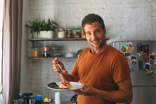 Close up portrait of a handsome young cheerful Caucasian man looking at camera while making classic tagliatelle by putting sauce on pasta on a plate in a kitchen.