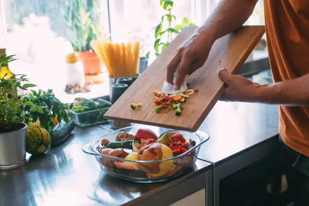 Close up shot of an anonymous young Caucasian man putting vegetable remains from a cutting board into a bowl on a kitchen counter.