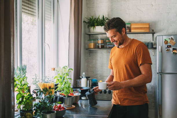 Handsome Young Man Holding a Coffee Maker and a Mug over a Kitchen Counter Close up photo of a handsome young happy Caucasian man holding a coffee maker and a mug over a kitchen counter. hot handsome man stock pictures, royalty-free photos & images