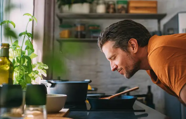 Handsome young happy Caucasian man enjoying the aroma of a meal with his eyes closed over a frying pan in a kitchen.