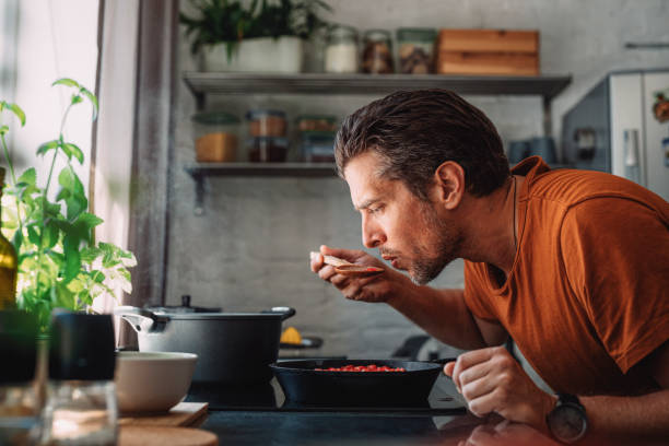 sauce de dégustation de jeune homme bel avec une cuillère de mélange dans une cuisine - cuisiner photos et images de collection