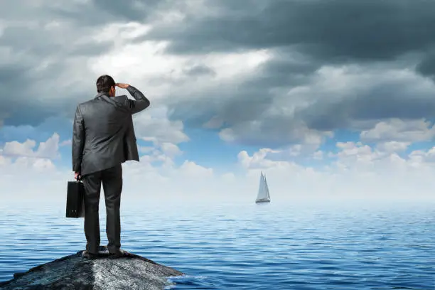 A man dressed in a suit and carrying a briefcase stands on a rock at the shore and looks out into the distance and realizes that his ship has sailed.