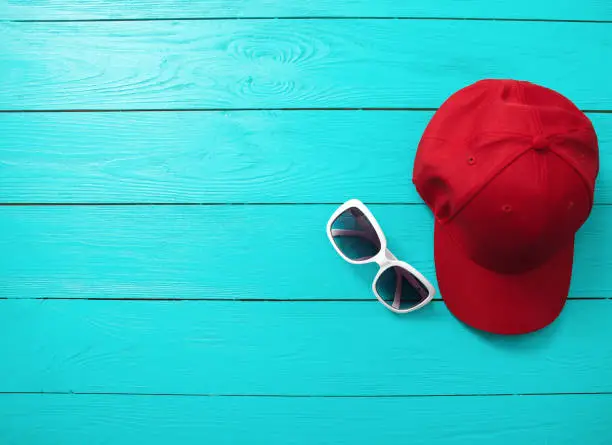 Photo of Red cap and sunglasses on blue wooden background. Top view and copy space. Summertime accessories.