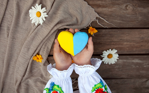 A yellow and blue heart in the hands of a child in an embroidered shirt, against the background of a rough wooden table and daisies.Unity Day, Independence Day of Ukraine, Embroidery Day