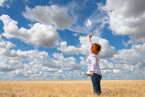 A young woman in a white shirt launches a paper airplane into the sky. A red-haired, cheerful girl enjoys life playing with a paper airplane. Concept: freedom and flight of the soul.