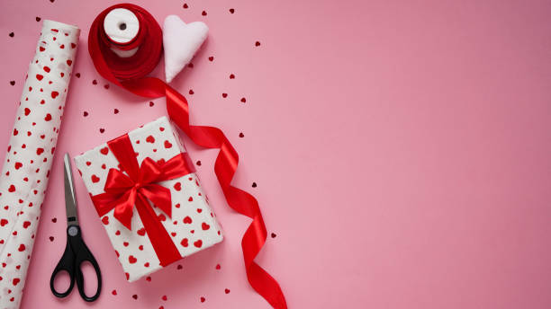 istock Valentine's day gift wrapping concept. Box wrapped in white paper in hearts,red bow, candy, red ribbon, reapers and a roll of wrapping paper on a pink background, top view, flat lay, copy space. 1297245311