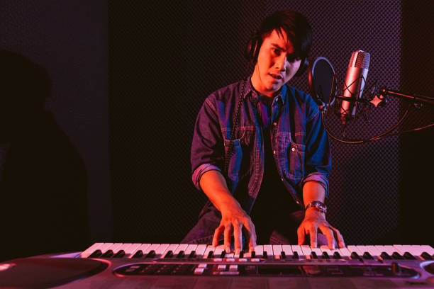 istock Male playing an electric keyboard while wearing headphones. Recording songs by using a studio microphone and pop shield on the microphone in blue and red light with copy space. 1297249967