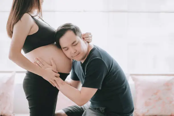 Photo of asian chinese man listening and leaning on his wife's abdomen with express love and anticipation for his lnew born