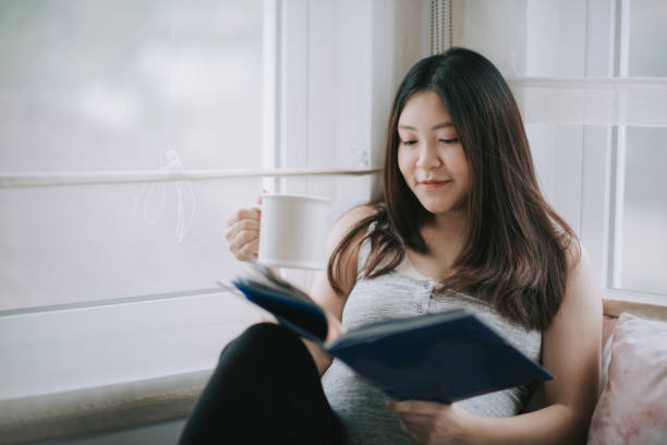 asian chinese pregnancy woman sitting on day time bed relaxing with a cup of tea enjoying reading book asian chinese pregnancy woman sitting on day time bed relaxing with a cup of tea enjoying reading book chaise longue woman stock pictures, royalty-free photos & images