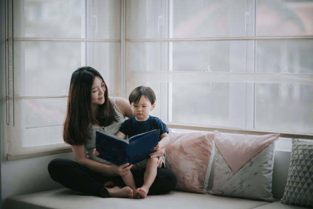 asian chinese pregnancy woman sitting on day time bed relaxing with a cup of tea enjoying reading book with her son - love growth time of day cheerful imagens e fotografias de stock