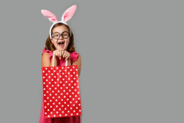 Photo of Happy easter. A cheerful little girl in a pink dress with polka dots holds a shopping bag in her hands. Cute baby on a gray background.
