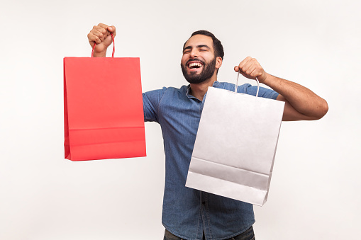Extremely happy satisfied man in blue shirt holding shopping paper bags, pleased with mall discounts, good purchases. Indoor studio shot isolated on white background