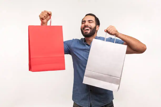 Photo of Extremely happy satisfied man in blue shirt holding shopping paper bags, pleased with mall discounts, good purchases