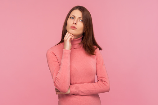 Thoughtful doubtful brunette woman standing holding her chin with hand, pondering about future, planning strategy. Indoor studio shot isolated on pink background