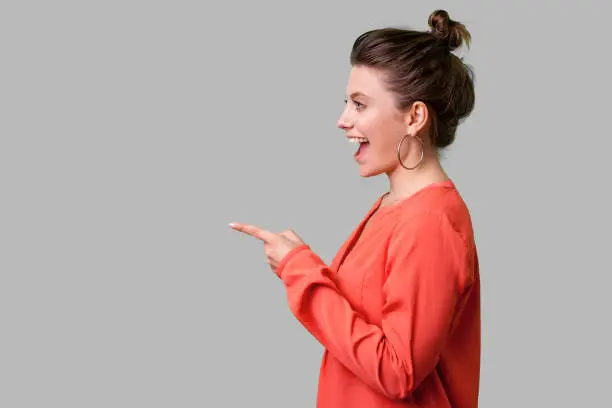 Hey you! Side view portrait of amazed girl with bun hairstyle, big earrings and in red blouse pointing finger to the left, looking with astonishment. indoor studio shot isolated on gray background