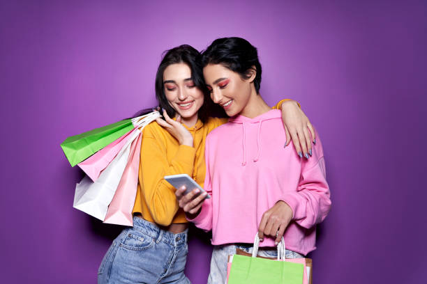 two happy women friends shoppers holding shopping bags using mobile apps for online shopping standing on purple background. retail ecommerce fashion sale offers, mall discounts in applications concept - 4369 imagens e fotografias de stock