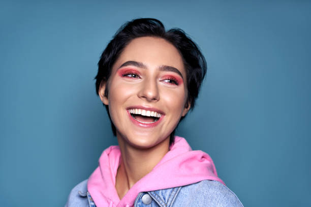 feliz cara adolescente con sonrisa amplia saludable dental y maquillaje rosa de moda riendo aislado en fondo azul. dientes blancos perfectos, odontología y cuidado de la piel, concepto cosmético adolescente. vista de primer plano - general fotografías e imágenes de stock