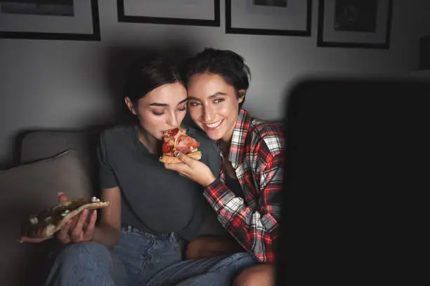 Photo of Two happy young girls eating pizza and watching tv at home party late. Best friends or couple having fun enjoying movie on television streaming service eating at night sitting on couch together.