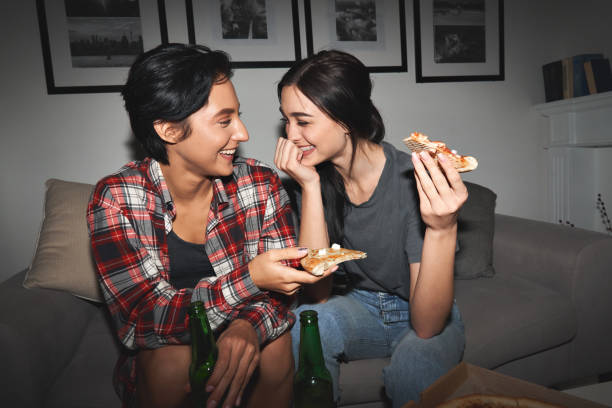 dwie szczęśliwe młode dziewczyny śmieją się, piją piwo, dzielą się pizzą na imprezie domowej późno. wesoły najlepsi przyjaciele lub para rozmawiają, bawiąc się jedząc fast food w nocy siedząc razem na kanapie. - pizza pizzeria friendship people zdjęcia i obrazy z banku zdjęć